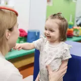 Uśmiechnięte dziecko w kwiecistym body wyciąga rękę do terapeuty w gabinecie.