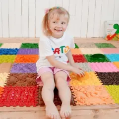 Uśmiechnięta dziewczynka bawi się na kolorowej macie sensorycznej dla niemowląt, odkrywając różne faktury i kształty.