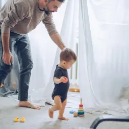 Ojciec wspiera dziecko, które stawia pierwsze kroki. Radość z momentu, kiedy dziecko zaczyna chodzić, jest widoczna na twarzy taty.