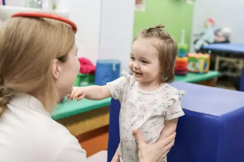 Uśmiechnięte dziecko w kwiecistym body wyciąga rękę do terapeuty w gabinecie.