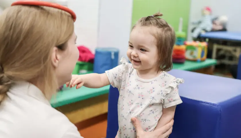 Uśmiechnięte dziecko w kwiecistym body wyciąga rękę do terapeuty w gabinecie.